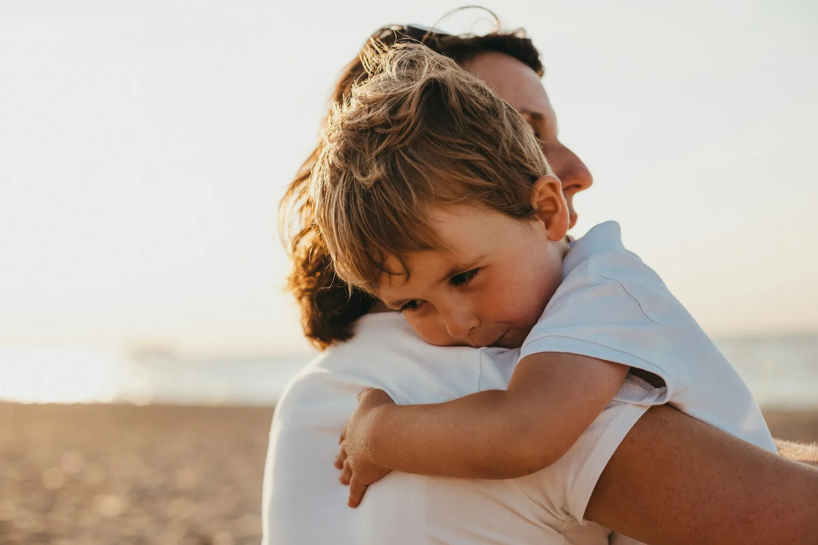 A mother holding her young child close on a sunlit beach.